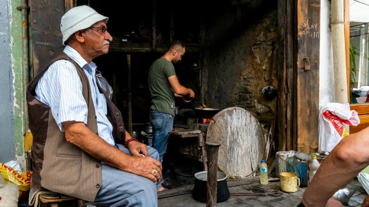 Elderly man sits outside a bustling blacksmith workshop, observing daily life on the street.