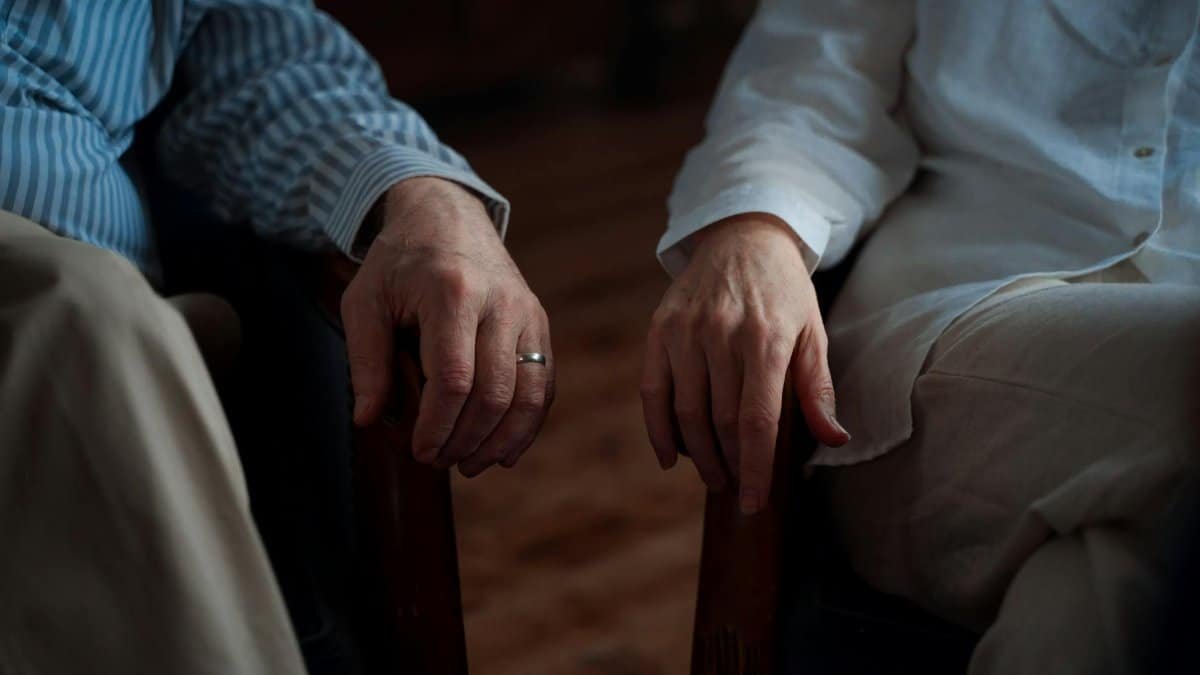 A touching moment capturing an elderly couple's hands resting on armchairs indoors.