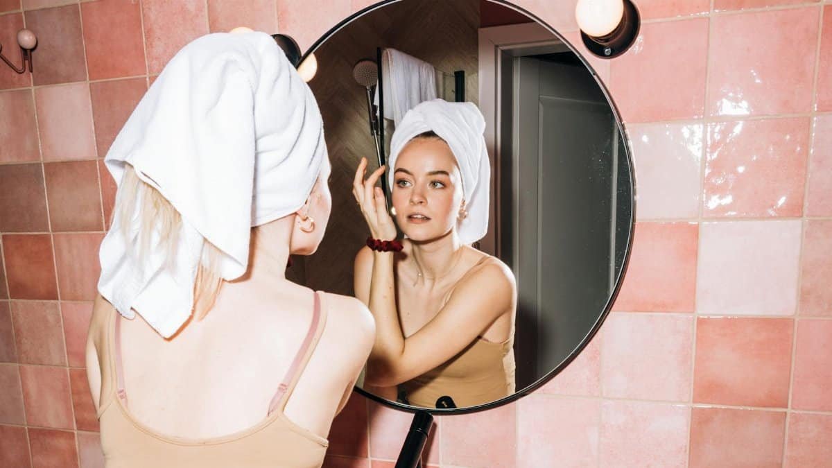 A young woman with a towel in a pink bathroom, reflecting in round mirror as she performs skincare.