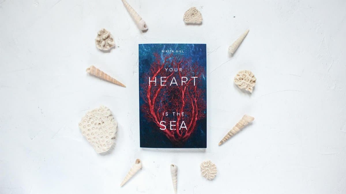 Flatlay featuring a book titled 'Your Heart is the Sea' surrounded by seashells on a white background.