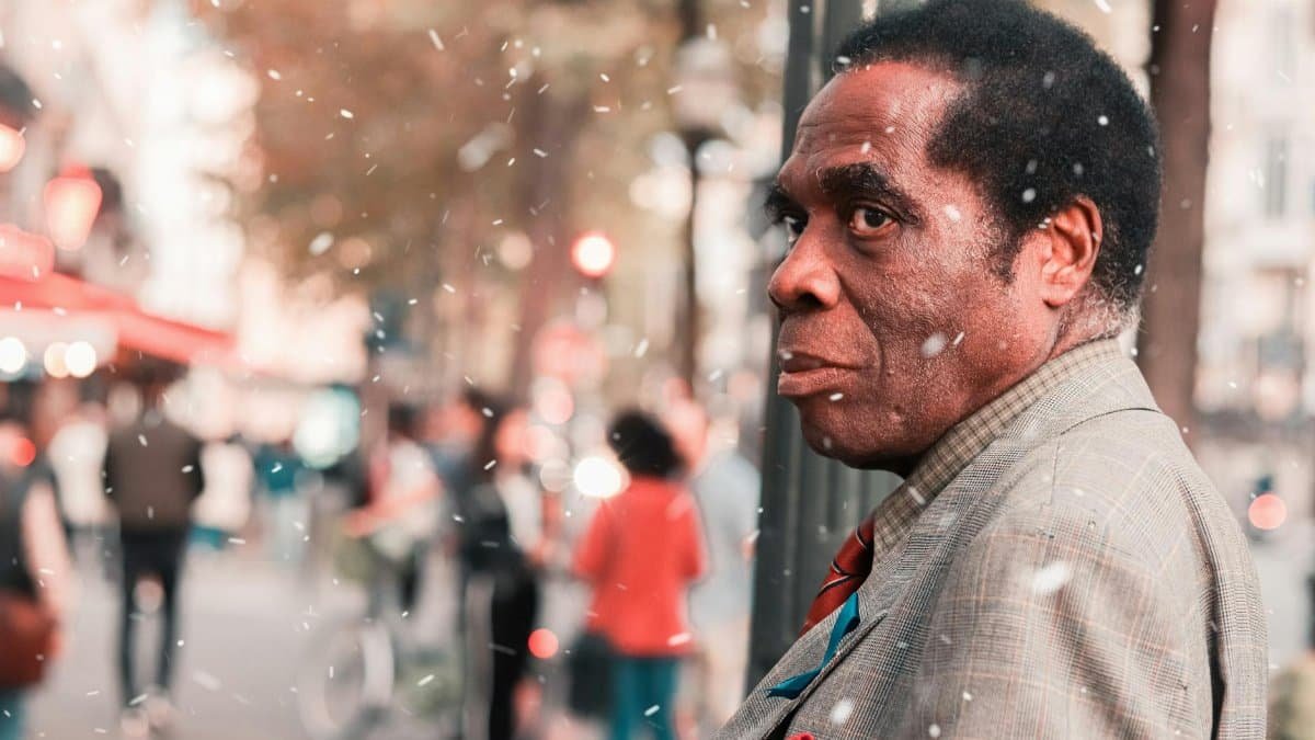 A man stands amidst a bustling city street under a gentle snowfall, captured in warm tones.