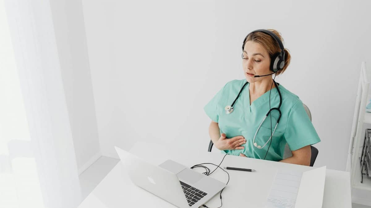 Medical professional using laptop for virtual healthcare session, wearing headset and scrubs.