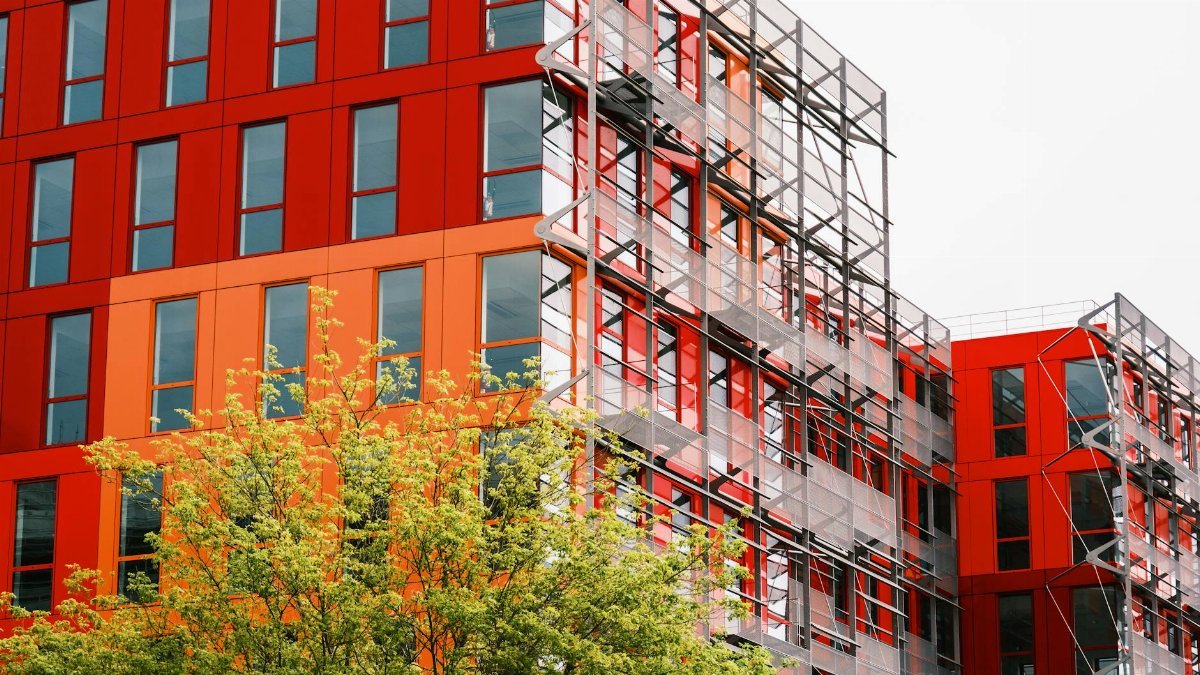 Striking red building facade with windows and scaffolding surrounded by greenery captures urban development.