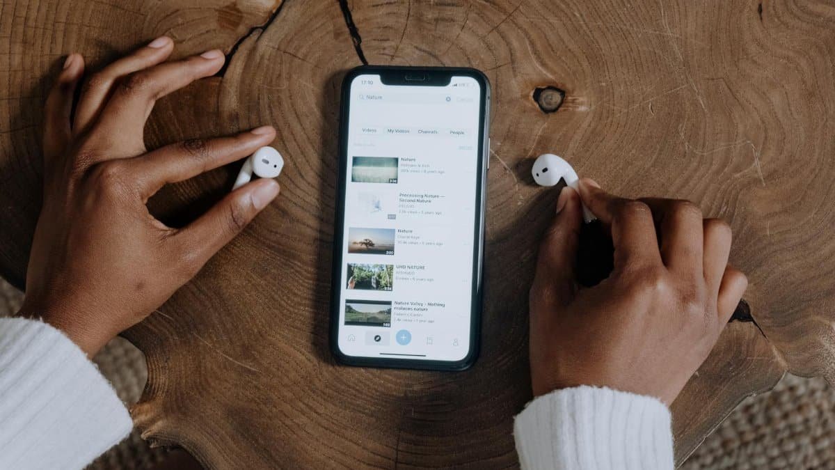 Hands interacting with smartphone and earbuds on a rustic wooden table, highlighting modern technology.