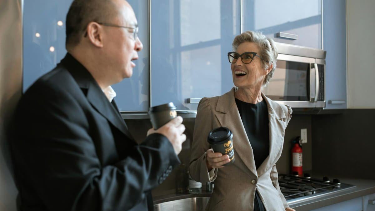 Two senior adults sharing a laugh over coffee in a modern office kitchen setting.