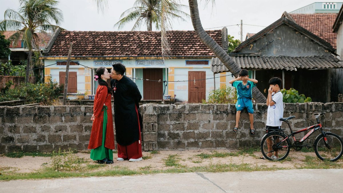 A couple in traditional attire kisses outdoors, with children observing from a stone wall.