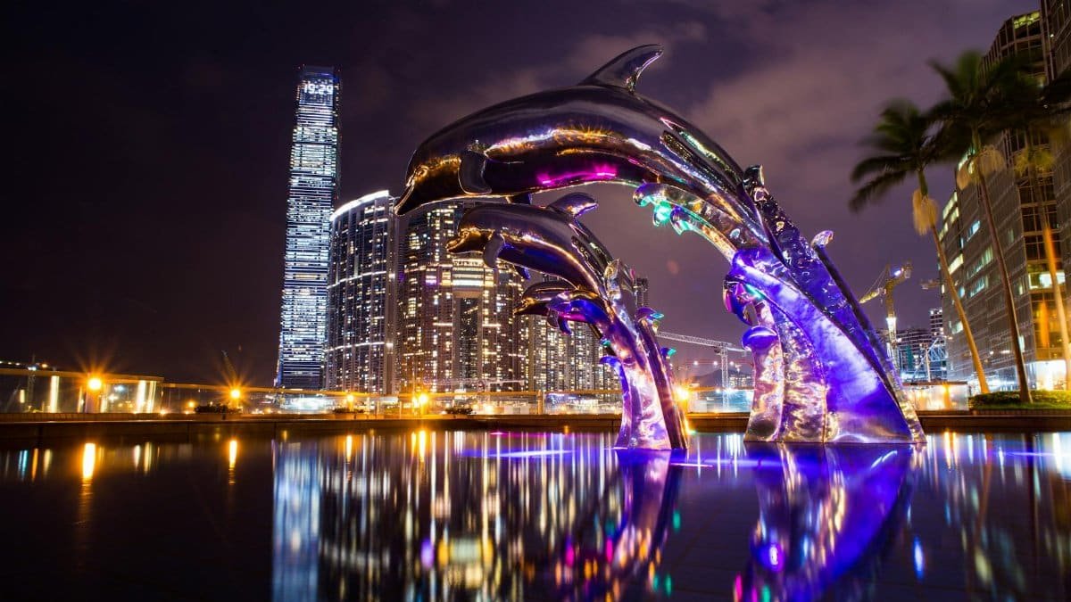 Dramatic cityscape at night featuring illuminated dolphin sculpture with reflections.