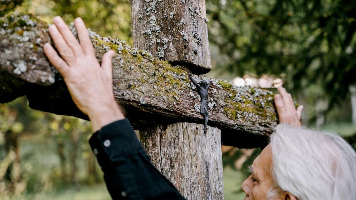 An elderly man reflects by a moss-covered wooden cross in a serene outdoor setting.