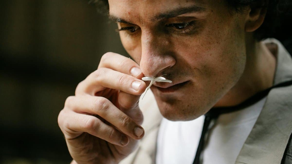 A man thoughtfully smells a delicate white flower in an outdoor setting, enjoying its scent.