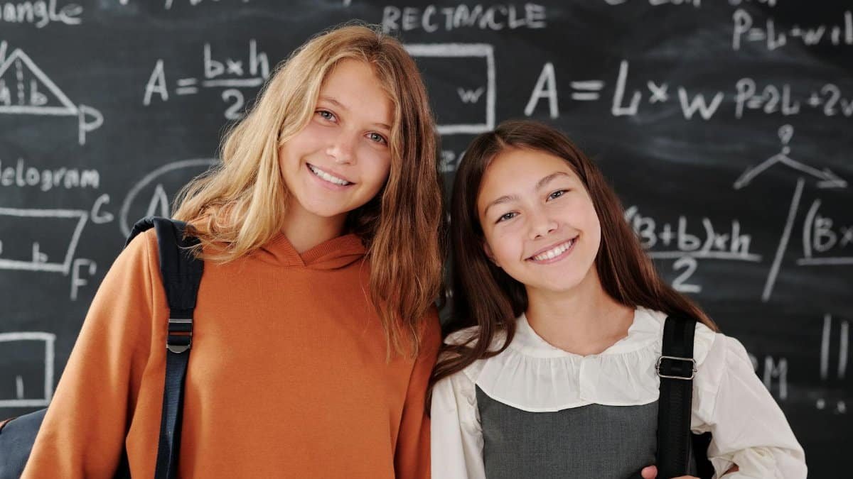 Two cheerful girls stand in a classroom with a math-covered chalkboard, ready for learning.