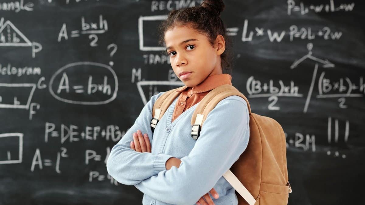 African American girl with backpack confidently poses in front of math-filled chalkboard in classroom.