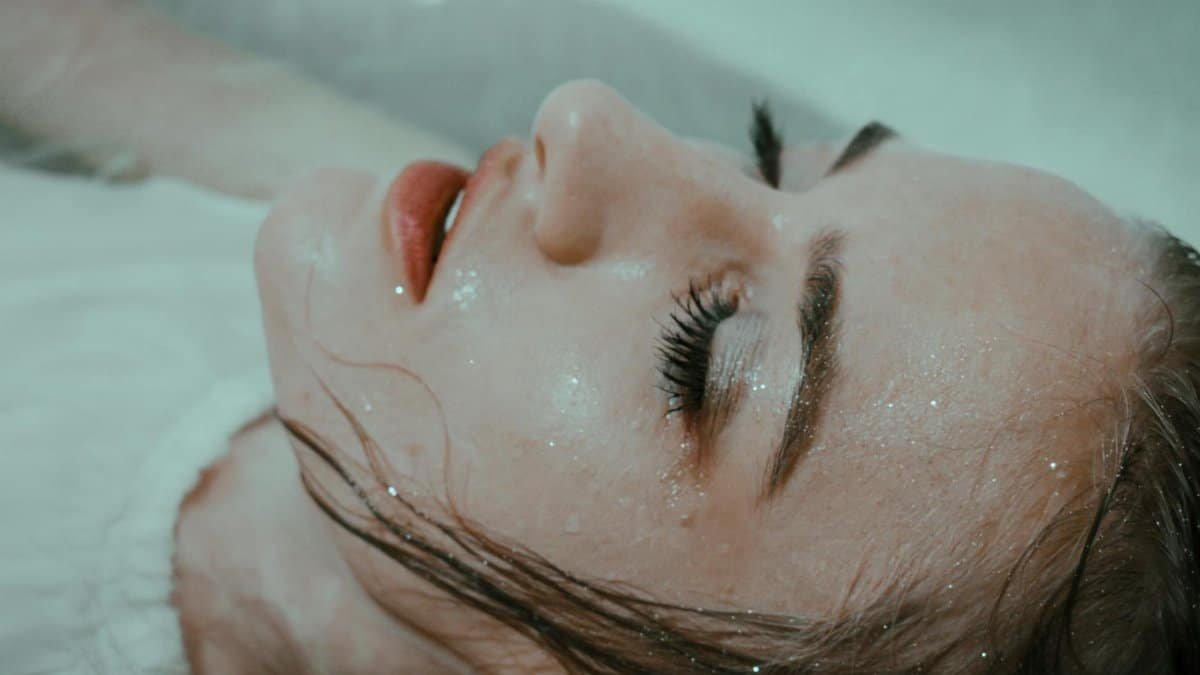 Relaxed female wearing white outfit with wet hair lying in bathtub in water with closed eyes at home in bathroom