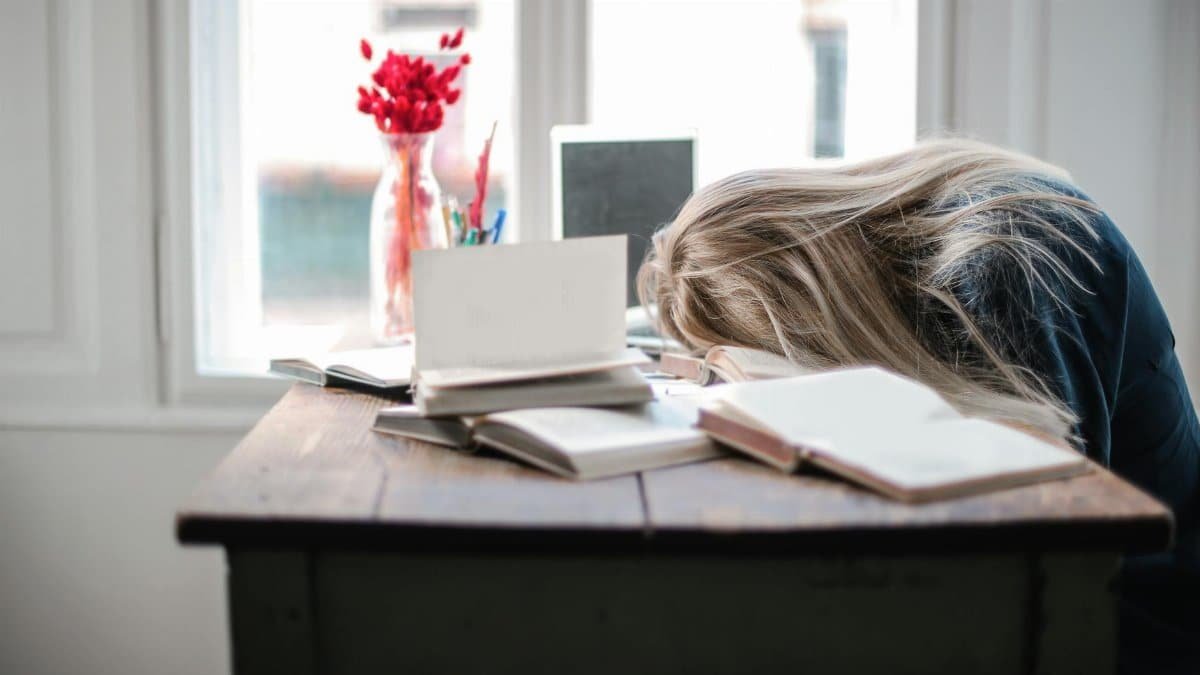 Blonde woman slumped over desk filled with books, showcasing study fatigue indoors.