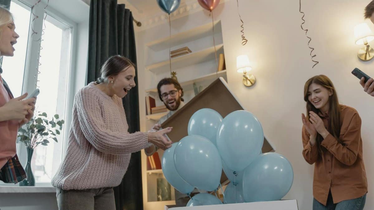 Friends joyfully celebrate a gender reveal with blue balloons indoors.