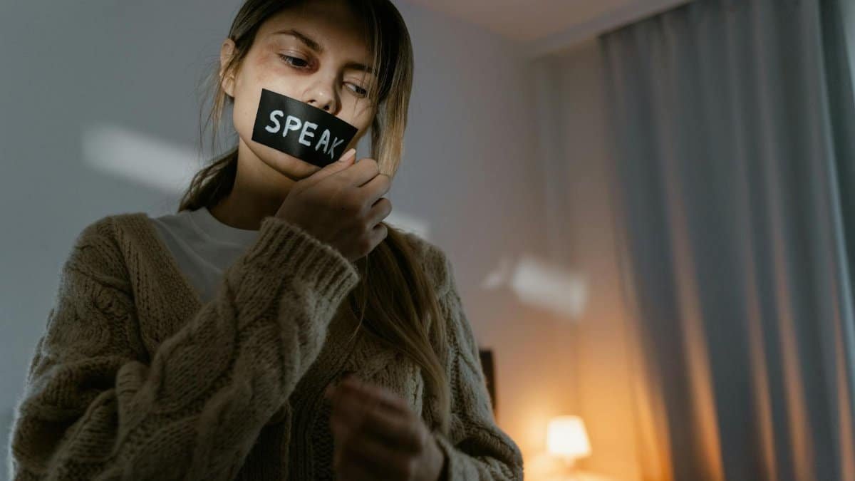 A woman standing in a dimly lit room removing tape with 'SPEAK' written, symbolizing empowerment.