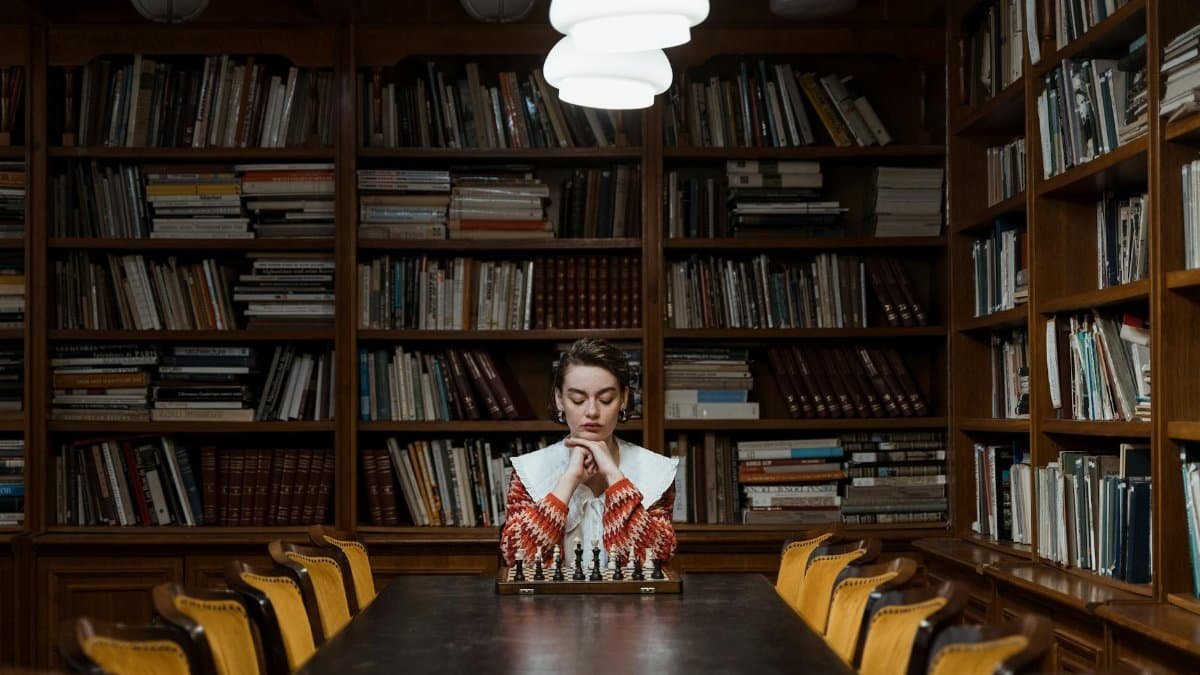 A focused woman ponders her chess move in a quiet library setting.