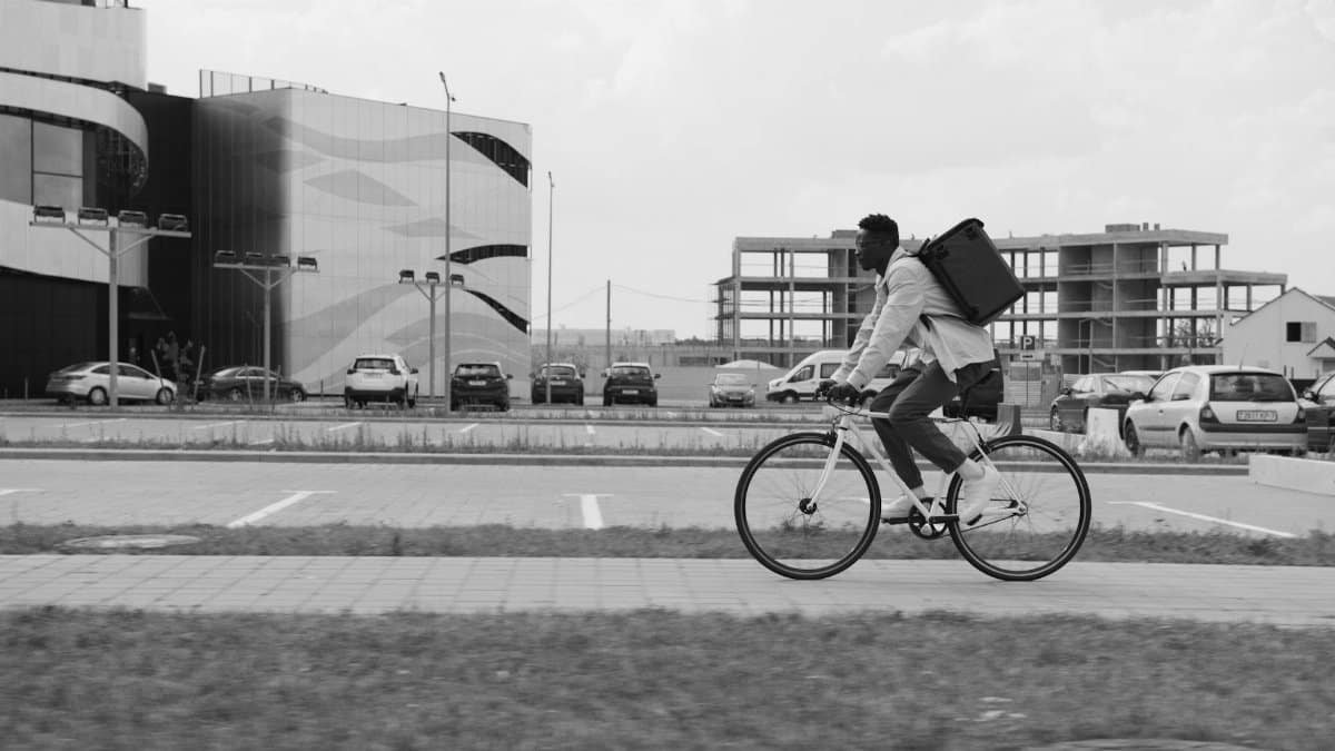 Black and white image of a deliveryman cycling on a city street with modern architecture.
