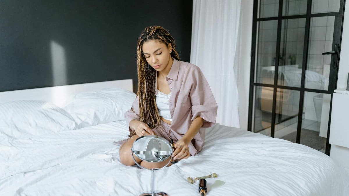 A woman with braided hair sits on a bed, focusing on self-care with a mirror.