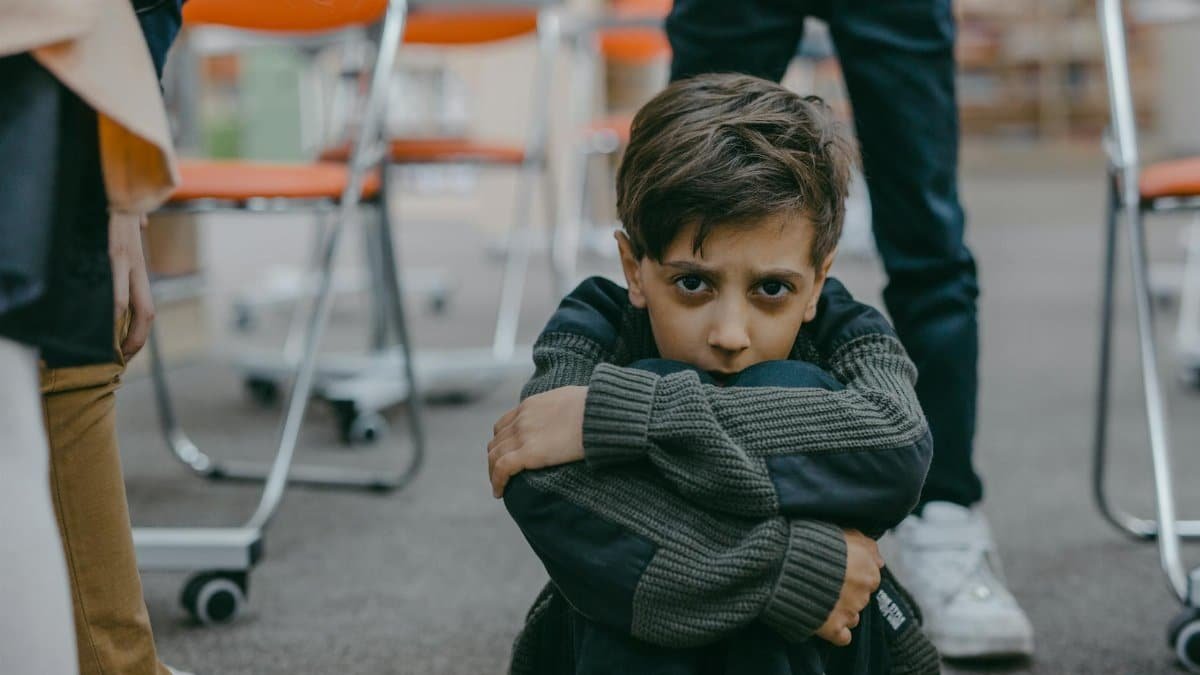 A young boy sits on the floor of a classroom, expressing fear and vulnerability.