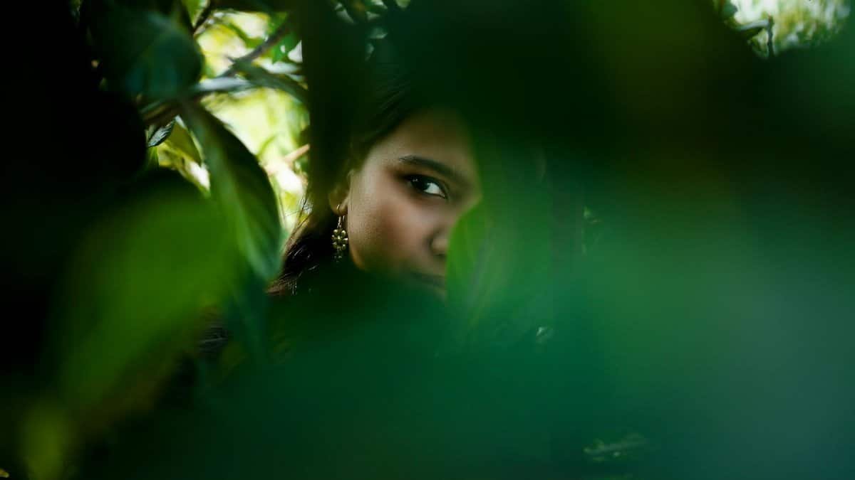 A woman partially hidden within lush green leaves, creating a sense of mystery.