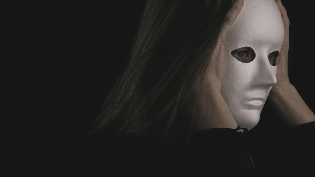 A person wearing a white mask held with both hands against a dark background, creating a mysterious atmosphere.