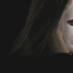 A person wearing a white mask held with both hands against a dark background, creating a mysterious atmosphere.