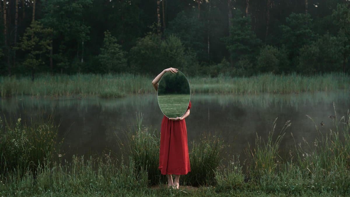 Artistic photo of a woman in nature holding a mirror beside a misty pond.