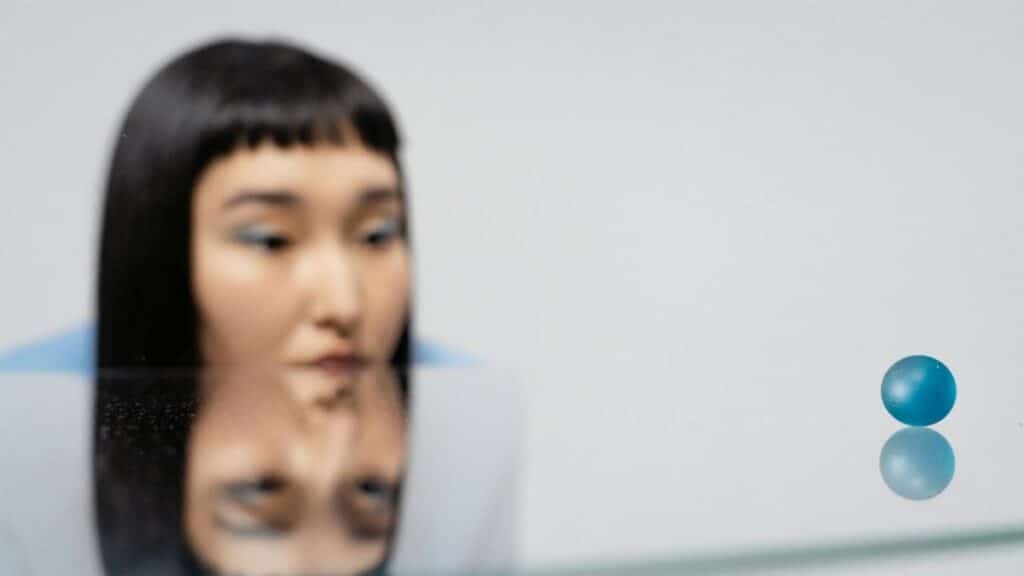 A woman gazes thoughtfully at a blue ball with reflections on a glass surface.