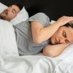 A woman looks frustrated in bed as her partner snores loudly, highlighting sleep disturbance issues.