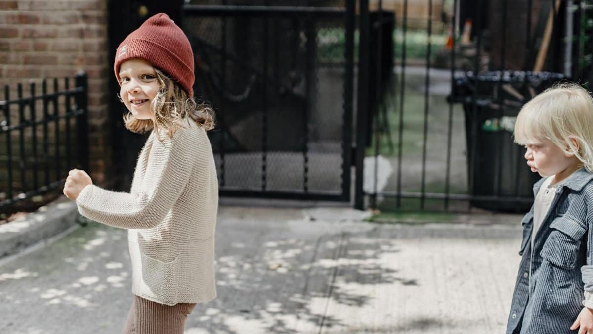 Side view of happy girl in hat and little boy strolling on sunny walkway near metal gates on street in city
