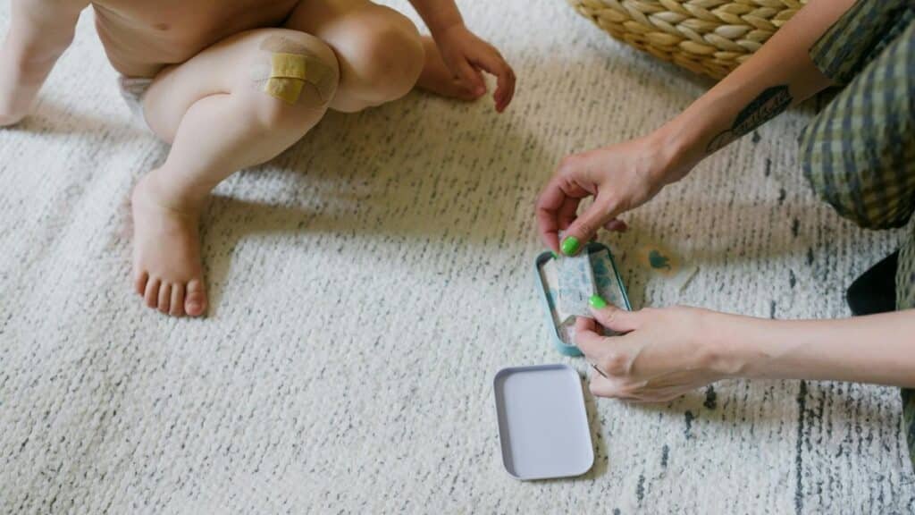 From above of crop anonymous barefooted little child with wound on knee sitting on carpet and choosing adhesive plaster with unrecognizable tattooed mother