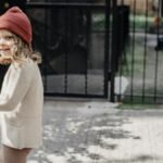 Side view of happy girl in hat and little boy strolling on sunny walkway near metal gates on street in city