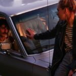 A dramatic moment between a couple by a classic car at dusk.