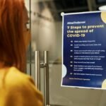 A woman reads a COVID-19 prevention poster emphasizing safety guidelines indoors.