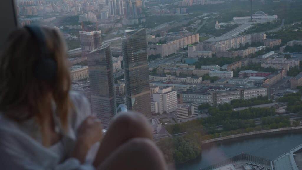 A woman wearing headphones views the cityscape from a high-rise window at sunset.