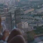 A woman wearing headphones views the cityscape from a high-rise window at sunset.