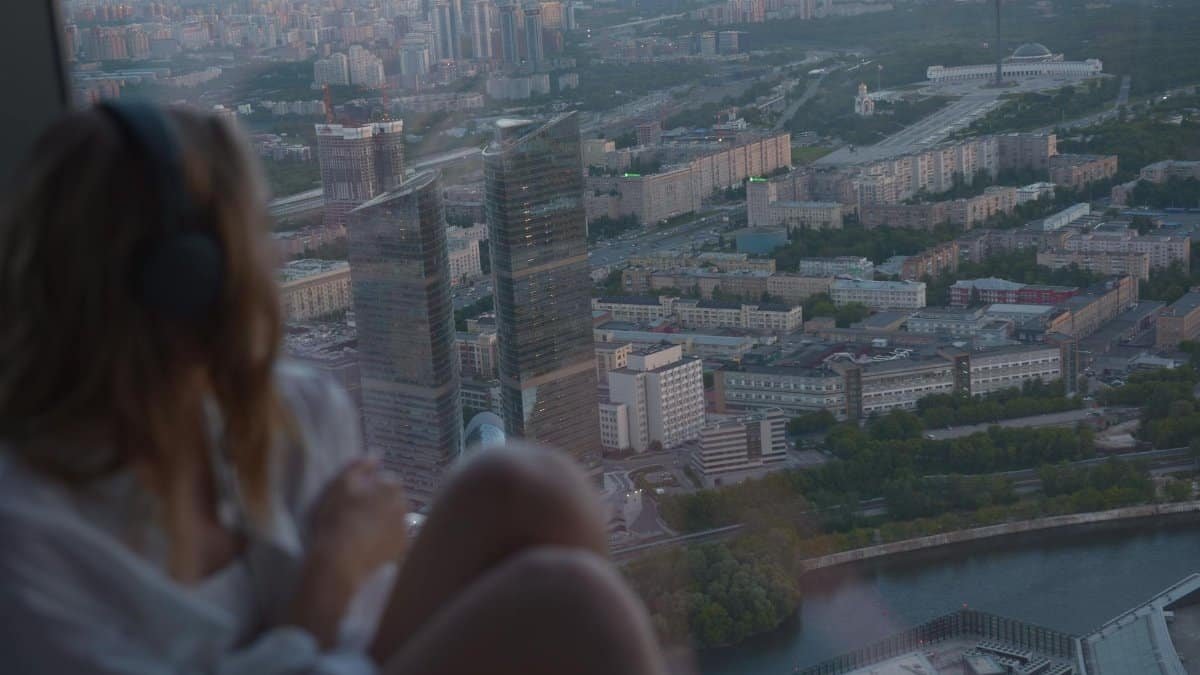 A woman wearing headphones views the cityscape from a high-rise window at sunset.