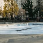 Deserted pool surrounded by high-rise buildings and autumn trees casting shadows in an urban setting.