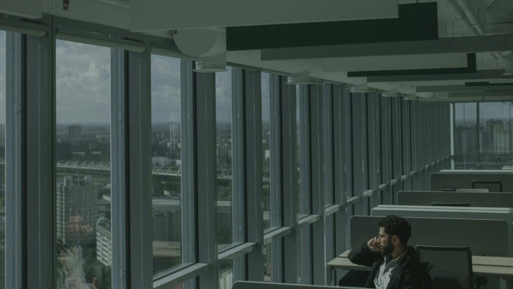 A man sits in a modern high-rise office, gazing thoughtfully through expansive windows at the city below.