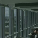 A man sits in a modern high-rise office, gazing thoughtfully through expansive windows at the city below.