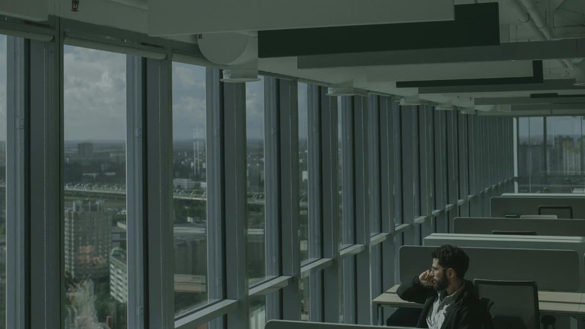 A man sits in a modern high-rise office, gazing thoughtfully through expansive windows at the city below.