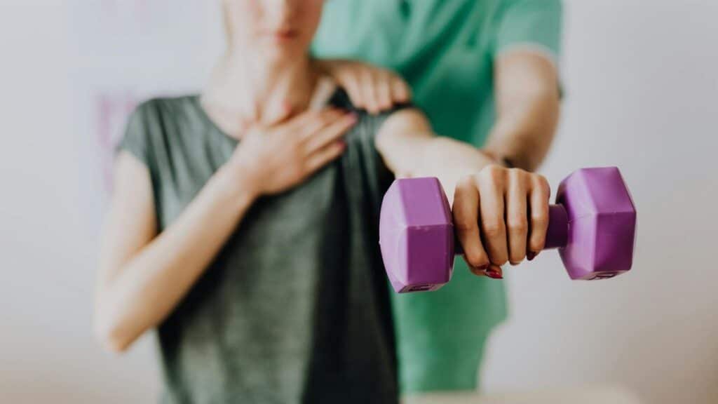 Crop unrecognizable orthopedist in uniform helping slender female in casual wear reaching arm with dumbbell in doctor office in hospital