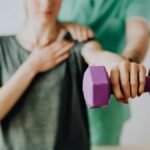 Crop unrecognizable orthopedist in uniform helping slender female in casual wear reaching arm with dumbbell in doctor office in hospital