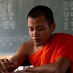 A focused Buddhist monk studying at a desk in an educational setting with a chalkboard in the background.