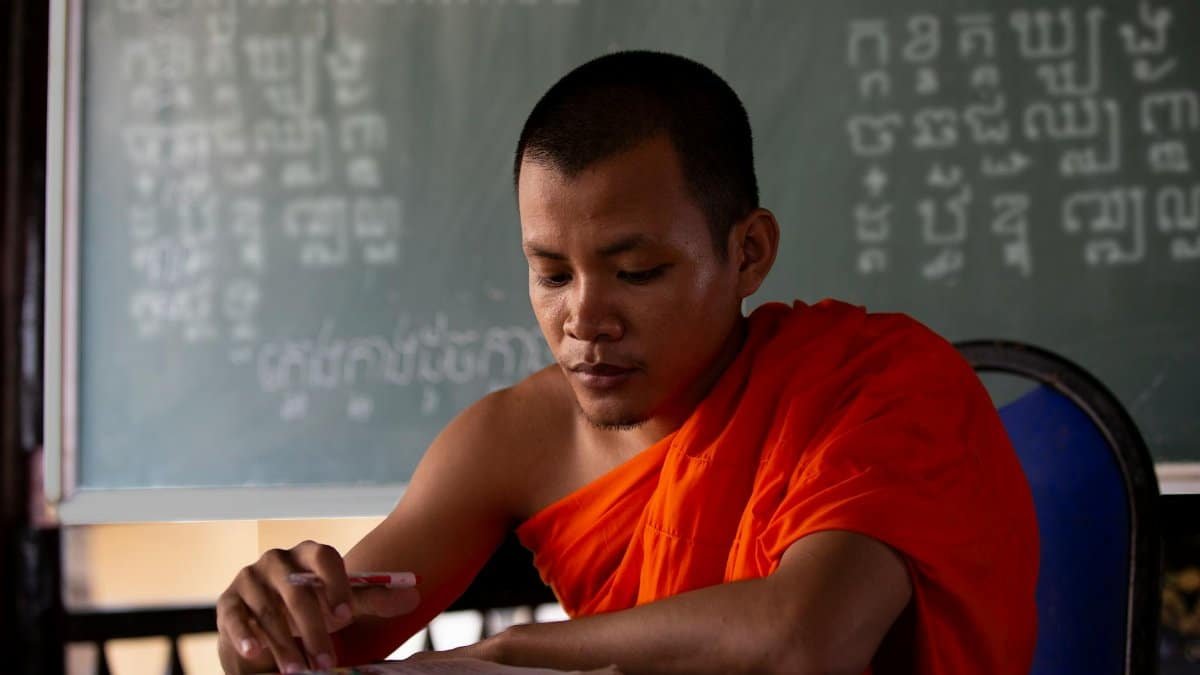 A focused Buddhist monk studying at a desk in an educational setting with a chalkboard in the background.