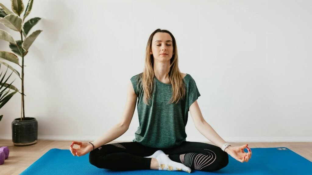 Serene blond lady in sportswear sitting with crossed legs and closed eyes while practicing yoga at home near potted plants