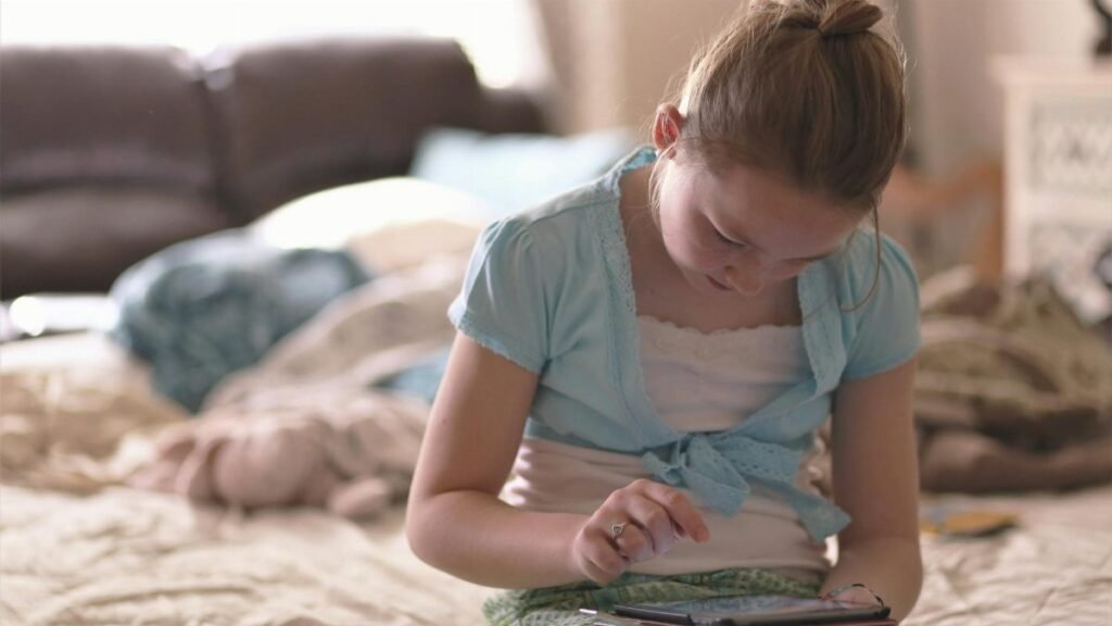 A young girl sitting on a bed, focused on using a tablet device in a cozy indoor setting.