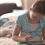 A young girl sitting on a bed, focused on using a tablet device in a cozy indoor setting.