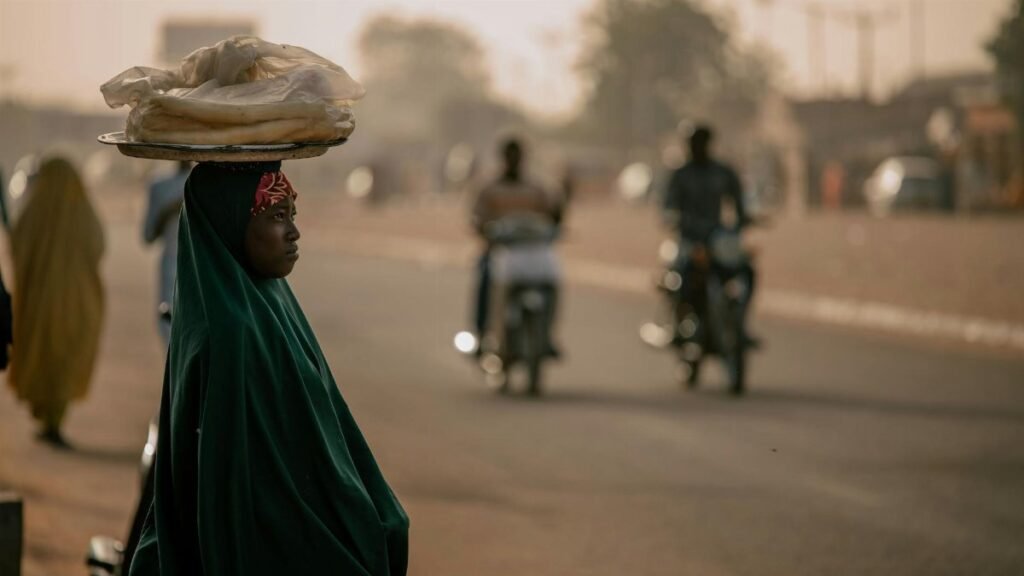 A woman balances a tray of bread on her head, walking along a busy street at sunset.