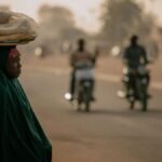 A woman balances a tray of bread on her head, walking along a busy street at sunset.